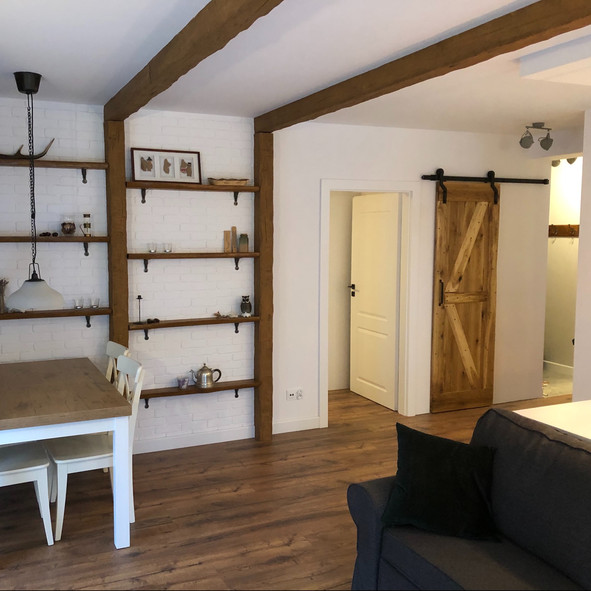 Jasne wnętrze z drewnianymi belkami stropowymi, ceglaną ścianą z półkami, przesuwnymi drzwiami stodołowymi i nowoczesnym oświetleniem.