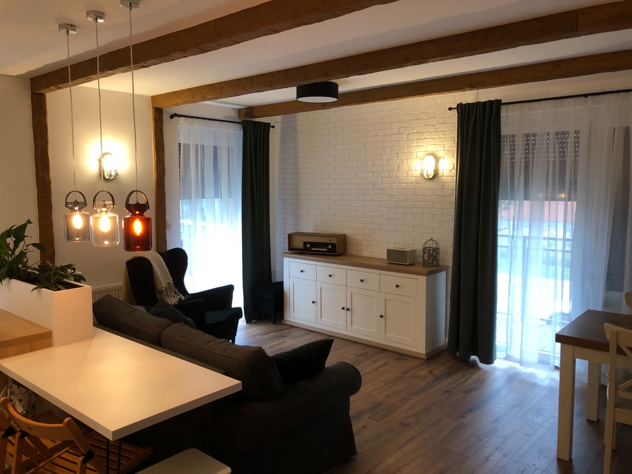 Jasne wnętrze salonu z drewnianymi belkami stropowymi, białymi cegłami na ścianie, ciemnymi zasłonami i parkietem, oświetlone trzema wiszącymi lampami w stylu retro.