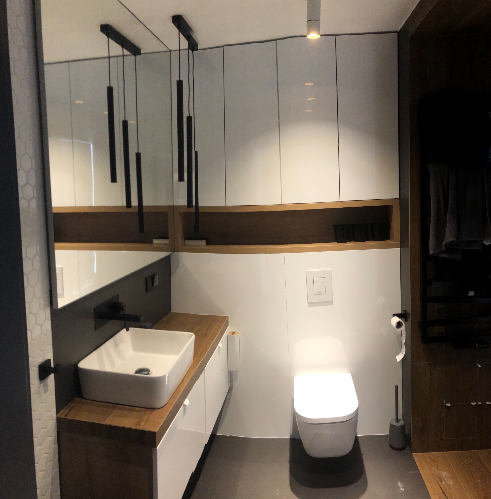 Nowoczesna łazienka z białą armaturą, drewnianym blatem pod umywalką i wiszącymi lampami w kształcie walców.