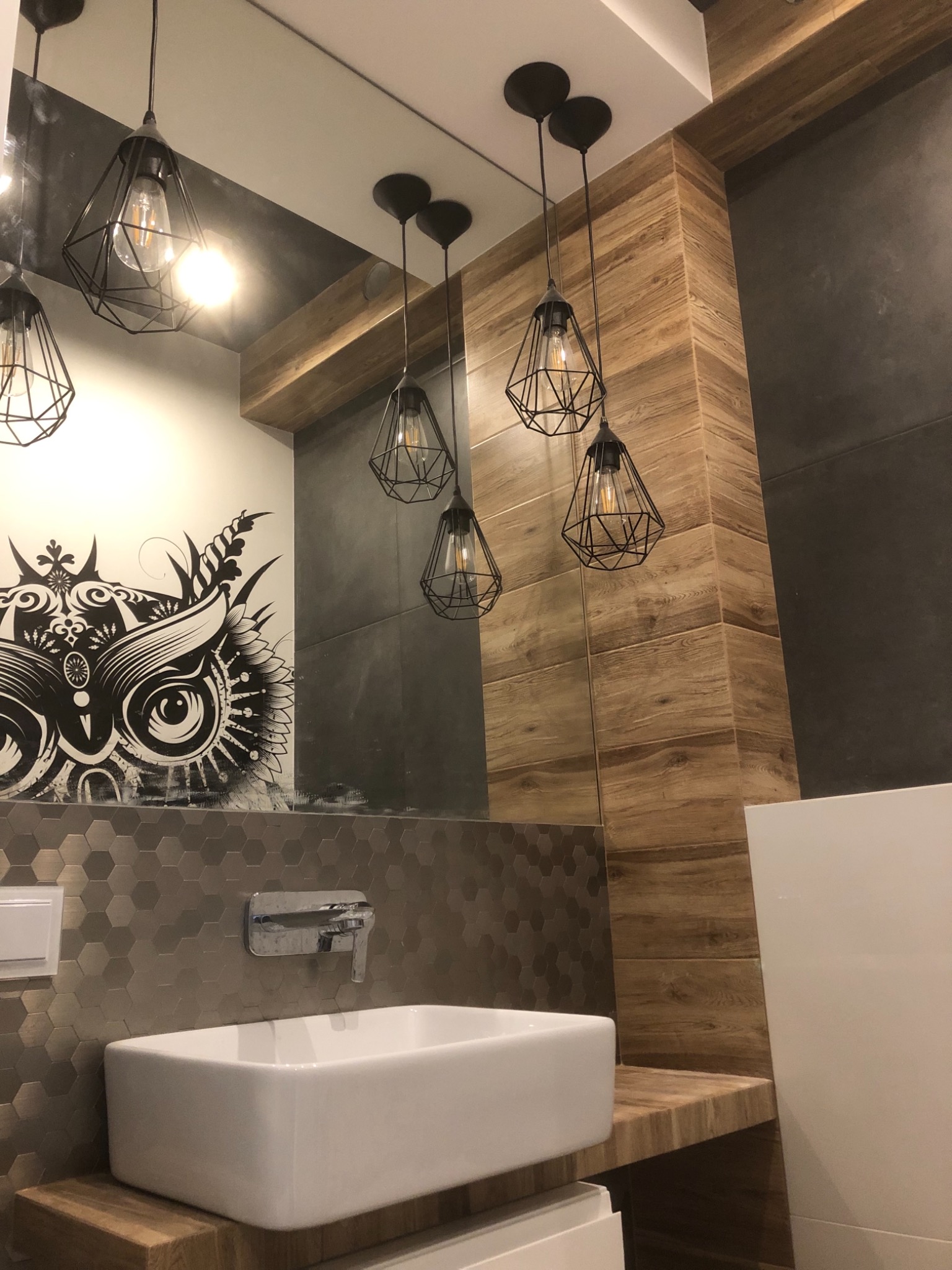 Nowoczesna łazienka z umywalką nablatową, heksagonalnymi płytkami, drewnopodobną okładziną ścienną i wiszącymi lampami w kształcie klatek, z odbiciem grafiki sowy w lustrze.