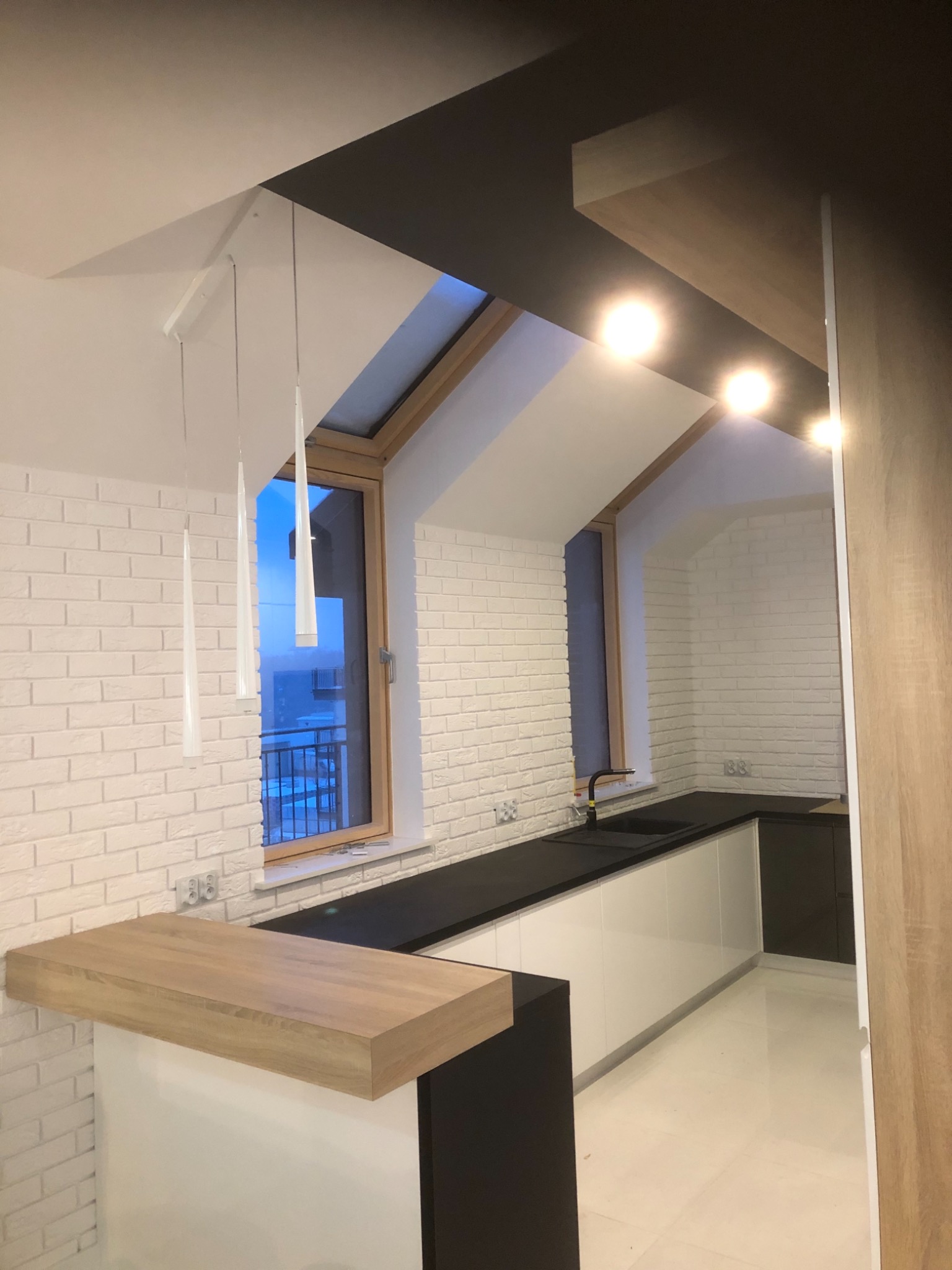 Nowoczesna kuchnia z białymi ceglanymi ścianami, czarnym blatem i białymi szafkami, oświetlona wiszącymi lampami i światłem wpadającym przez okno dachowe.