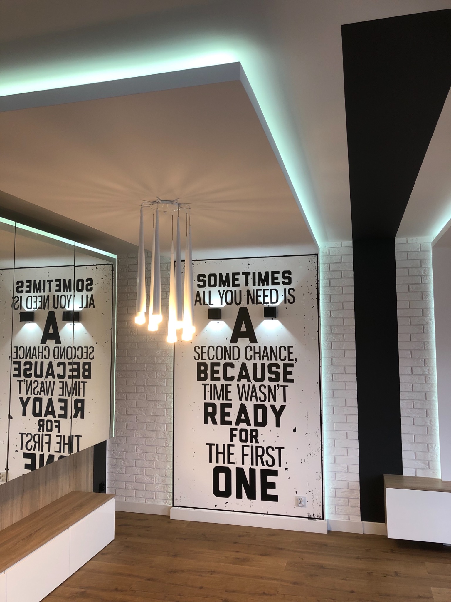 Nowoczesne oświetlenie sufitowe z podświetleniem LED, wisząca lampa z sześcioma białymi kloszami, ściana z białej cegły z czarnym pasem, dekoracyjny plakat z napisem, parkiet na podłodze.