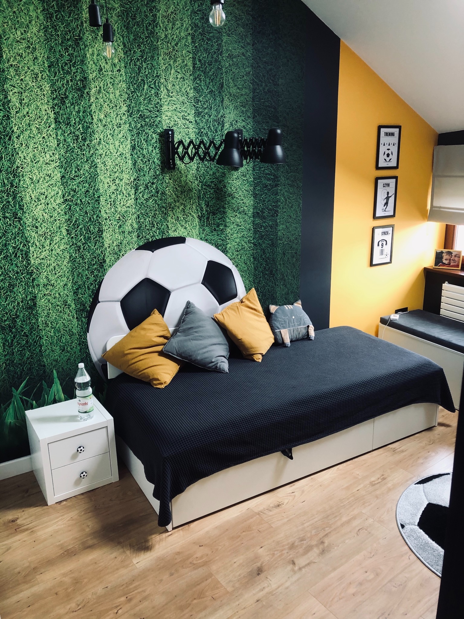 Pokój dziecięcy z łóżkiem z zagłówkiem w kształcie piłki, tapetą imitującą trawę boiska, czarną lampą harmonijkową, żółtą ścianą z ramkami i dywanem w kształcie piłki na podłodze z paneli.