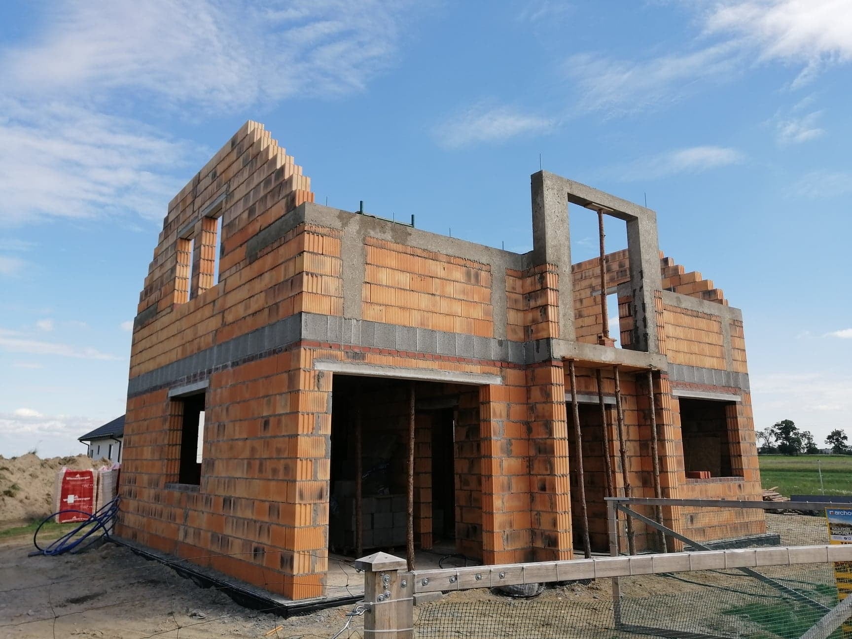 Budowa domu z czerwonej cegły ceramicznej w stanie surowym otwartym, widoczne otwory okienne i drzwiowe, szare betonowe nadproża i wieńce, błękitne niebo w tle.
