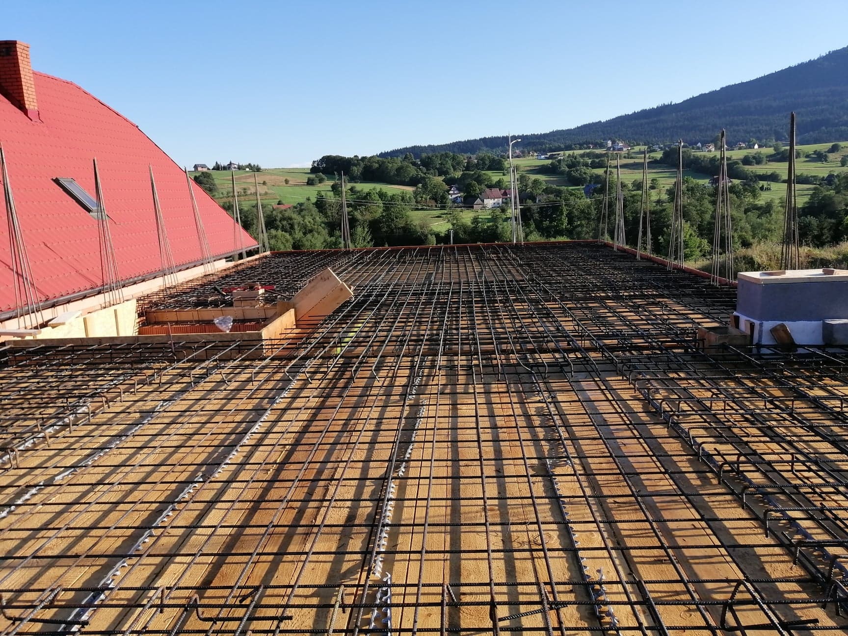 Zbrojenie płyty stropowej na drewnianym szalunku, widok na czerwoną dachówkę i malowniczy krajobraz górski w tle.