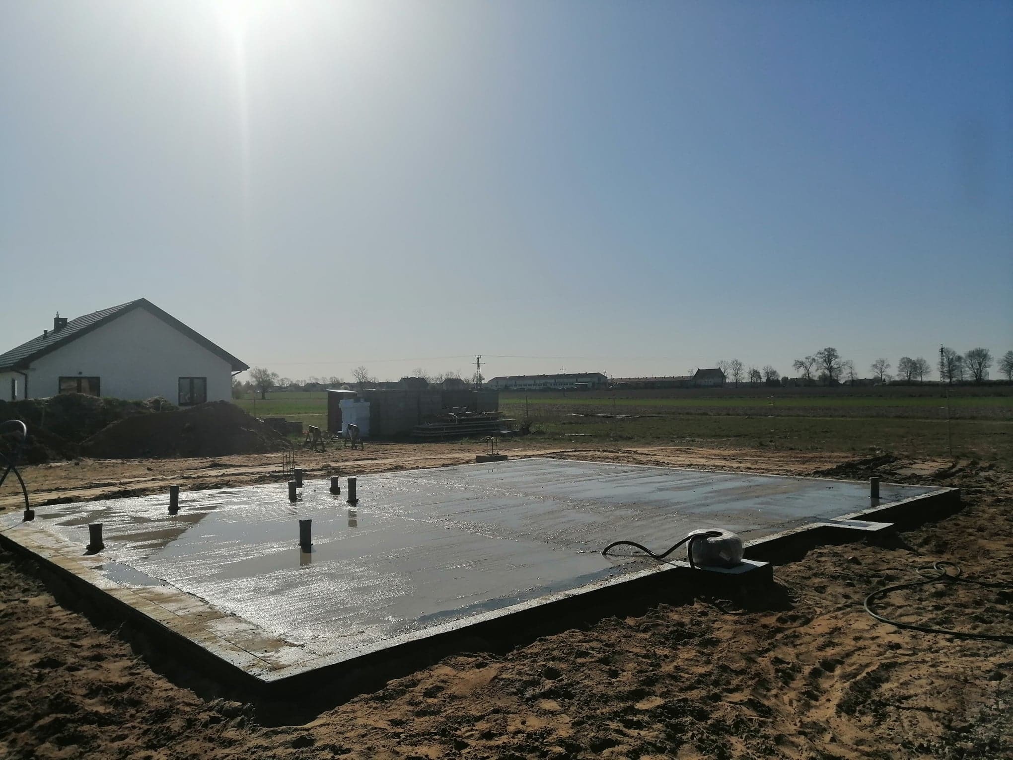 Świeżo wylana, mokra płyta fundamentowa z wystającymi rurami na działce budowlanej w słoneczny dzień, w tle widoczny dom jednorodzinny.