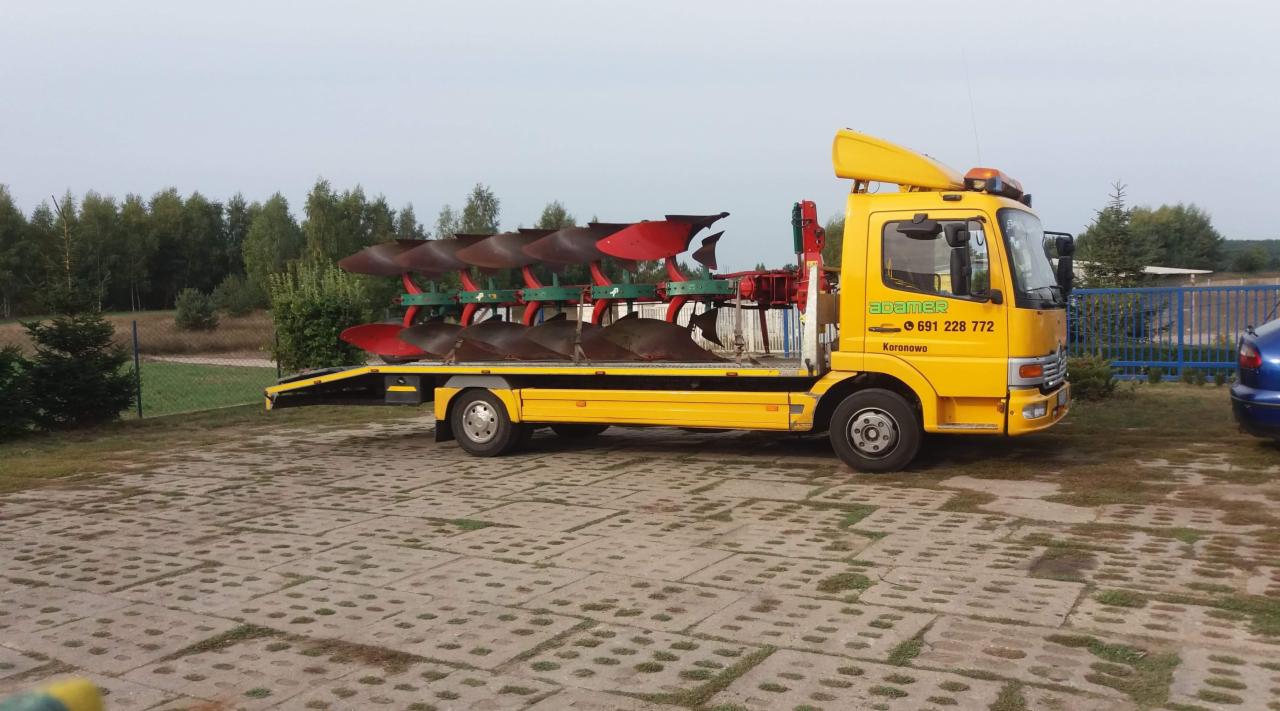 Żółta laweta z platformą transportową przewozi pług rolniczy z czerwonymi elementami na tle drzew i kostki brukowej.