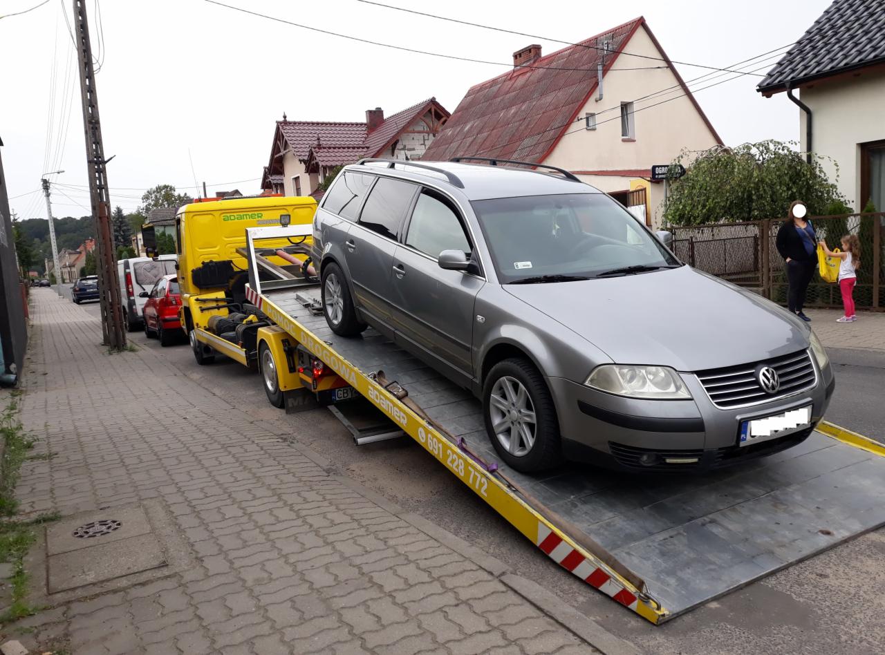 Szary samochód osobowy kombi na żółtej platformie pomocy drogowej z numerem telefonu, na tle domów i chodnika.