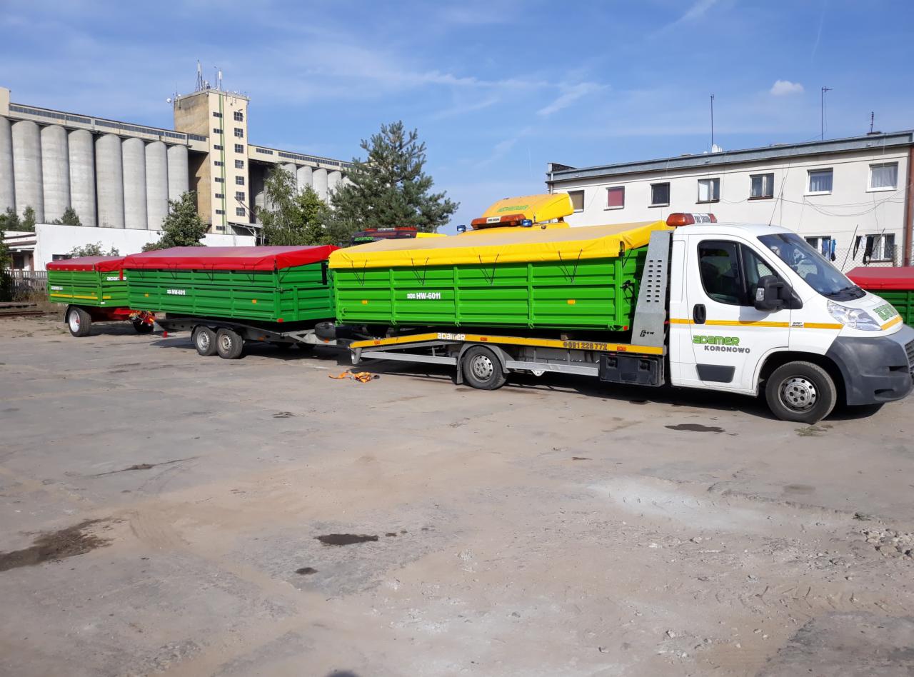 Biała laweta z żółtą plandeką transportuje dwa zielone przyczepy z czerwonymi i żółtymi plandekami na tle silosów zbożowych.