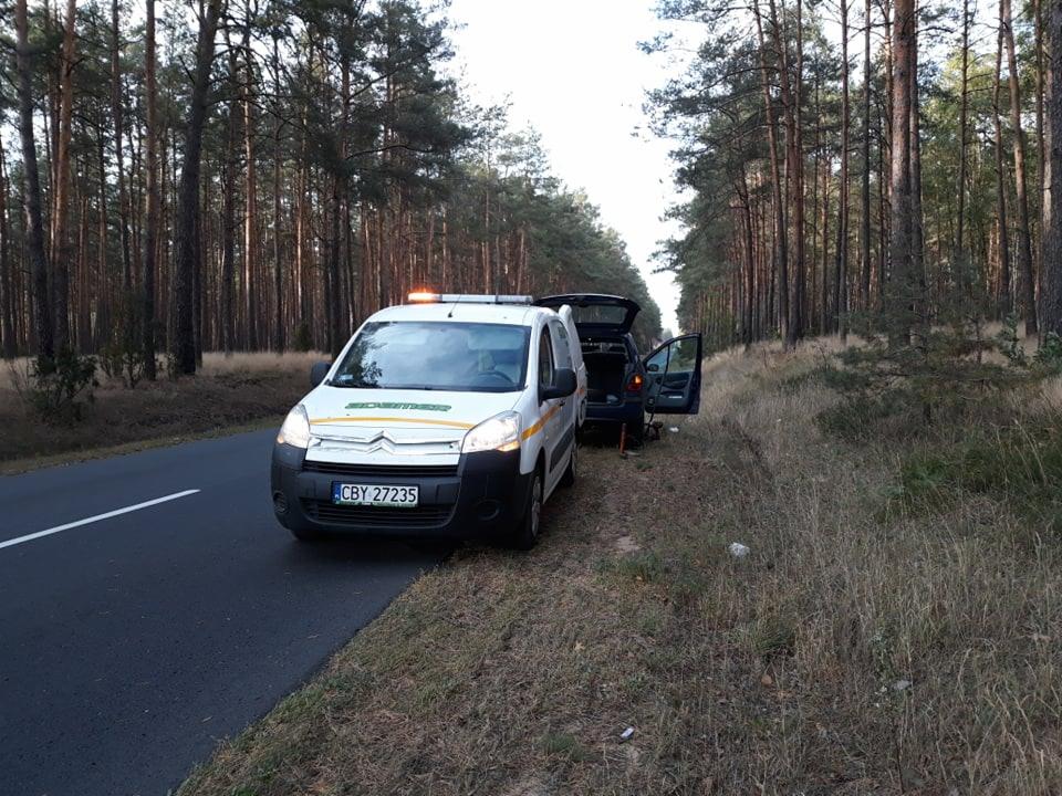 Biały bus pomocy drogowej z otwartym bagażnikiem i włączonym pomarańczowym światłem ostrzegawczym stoi na poboczu drogi w lesie, obok zaparkowany samochód osobowy z otwartymi drzwiami.