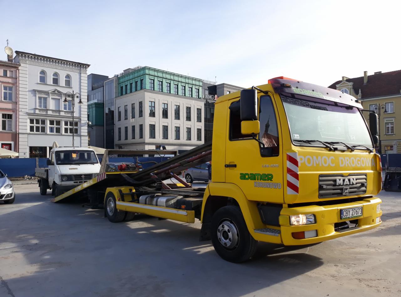 Żółta laweta MAN z podniesioną platformą, przewożąca biały samochód dostawczy na tle miejskiej zabudowy.