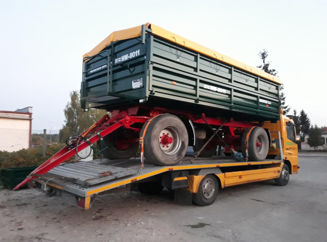 Żółta laweta przewozi zieloną przyczepę rolniczą z żółtą plandeką, model HW-8011, na tle zabudowań i drzew.