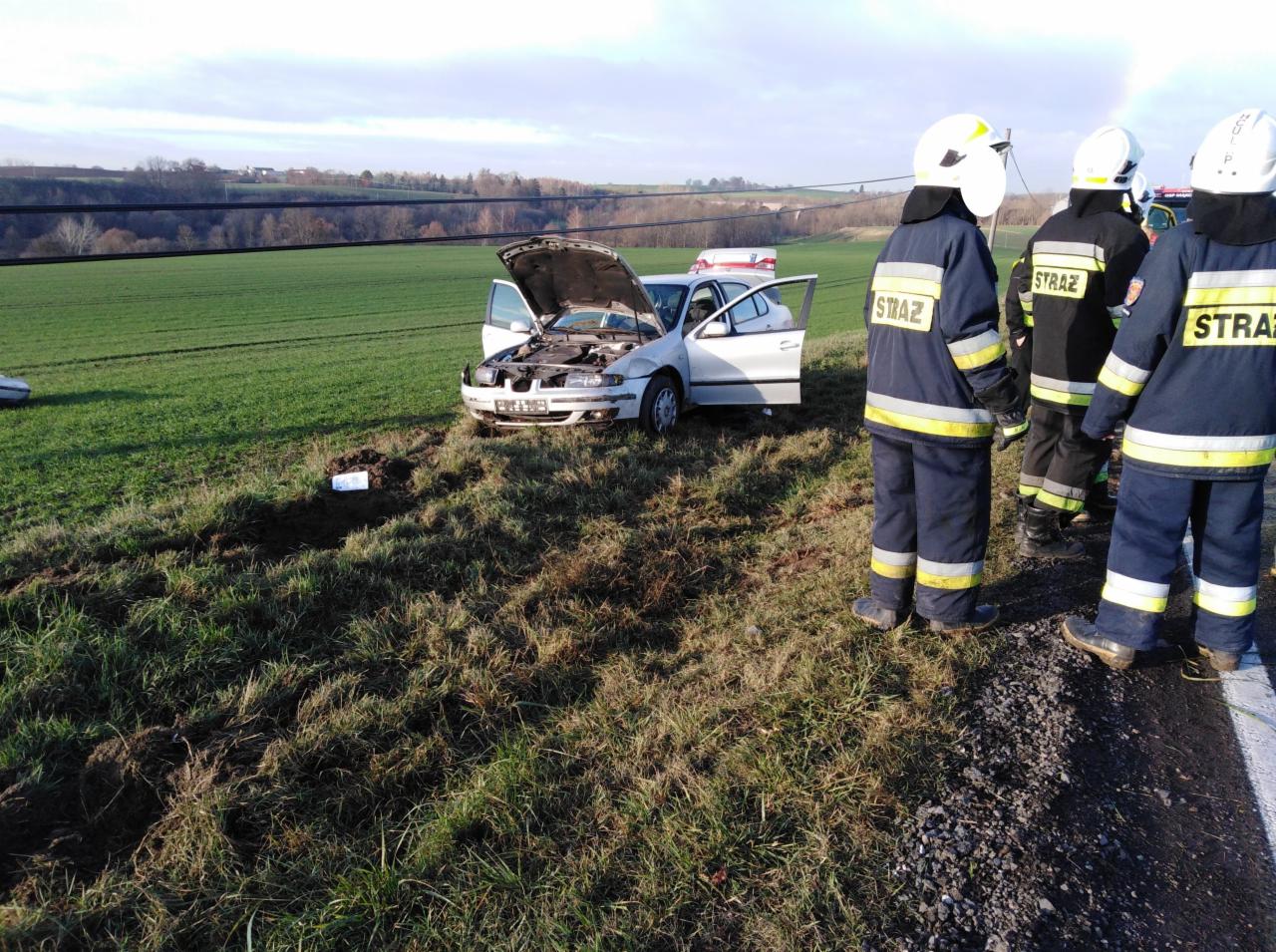 Uszkodzony srebrny samochód osobowy na poboczu pola, z otwartą maską i drzwiami, w tle strażacy w umundurowaniu, prawdopodobnie po wypadku.