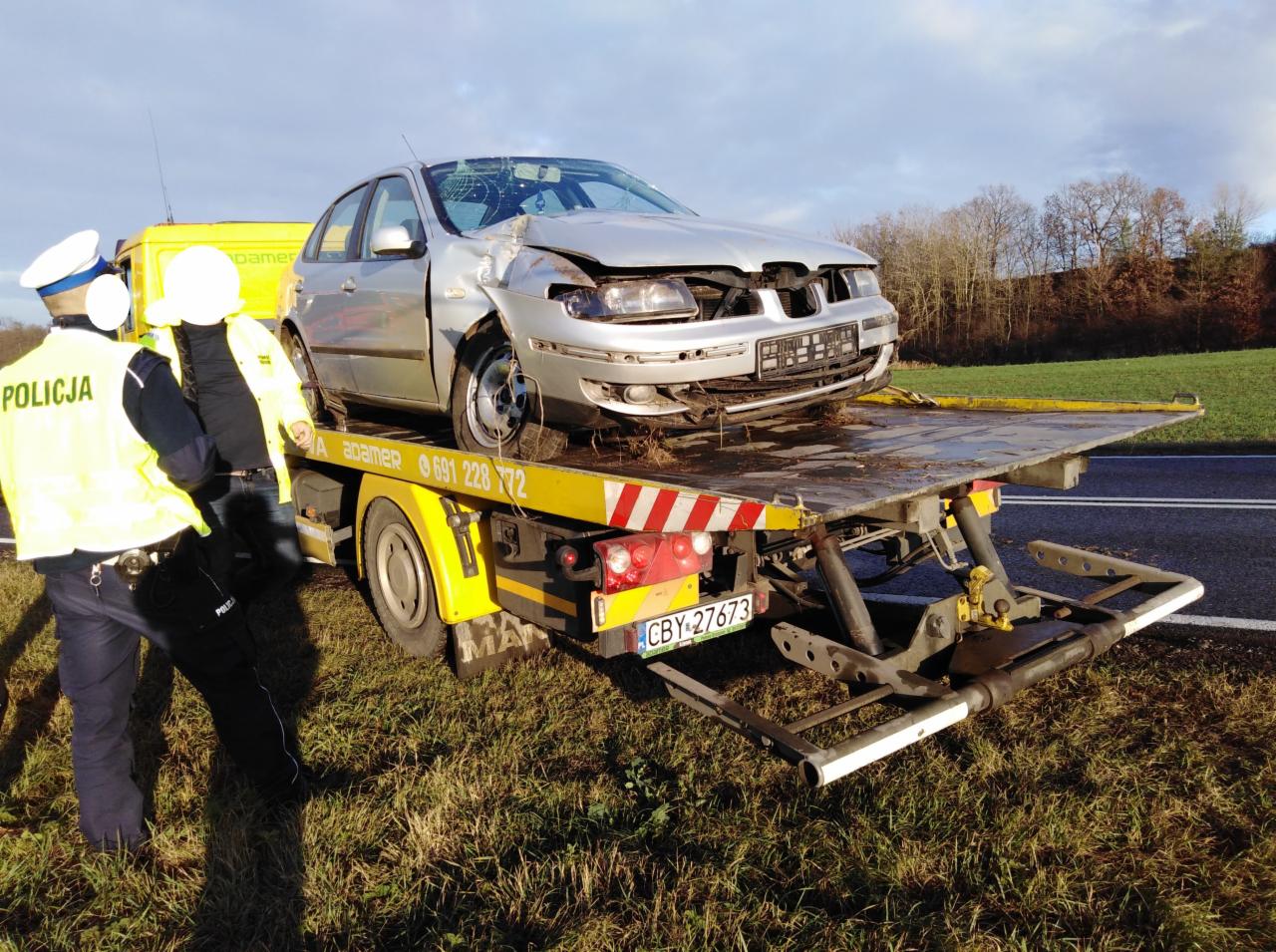 Uszkodzony srebrny samochód osobowy na platformie lawety, obok dwóch policjantów w mundurach. Widoczne zniszczenia przodu pojazdu i rozbita szyba. Laweta z żółtymi elementami i numerem telefonu.