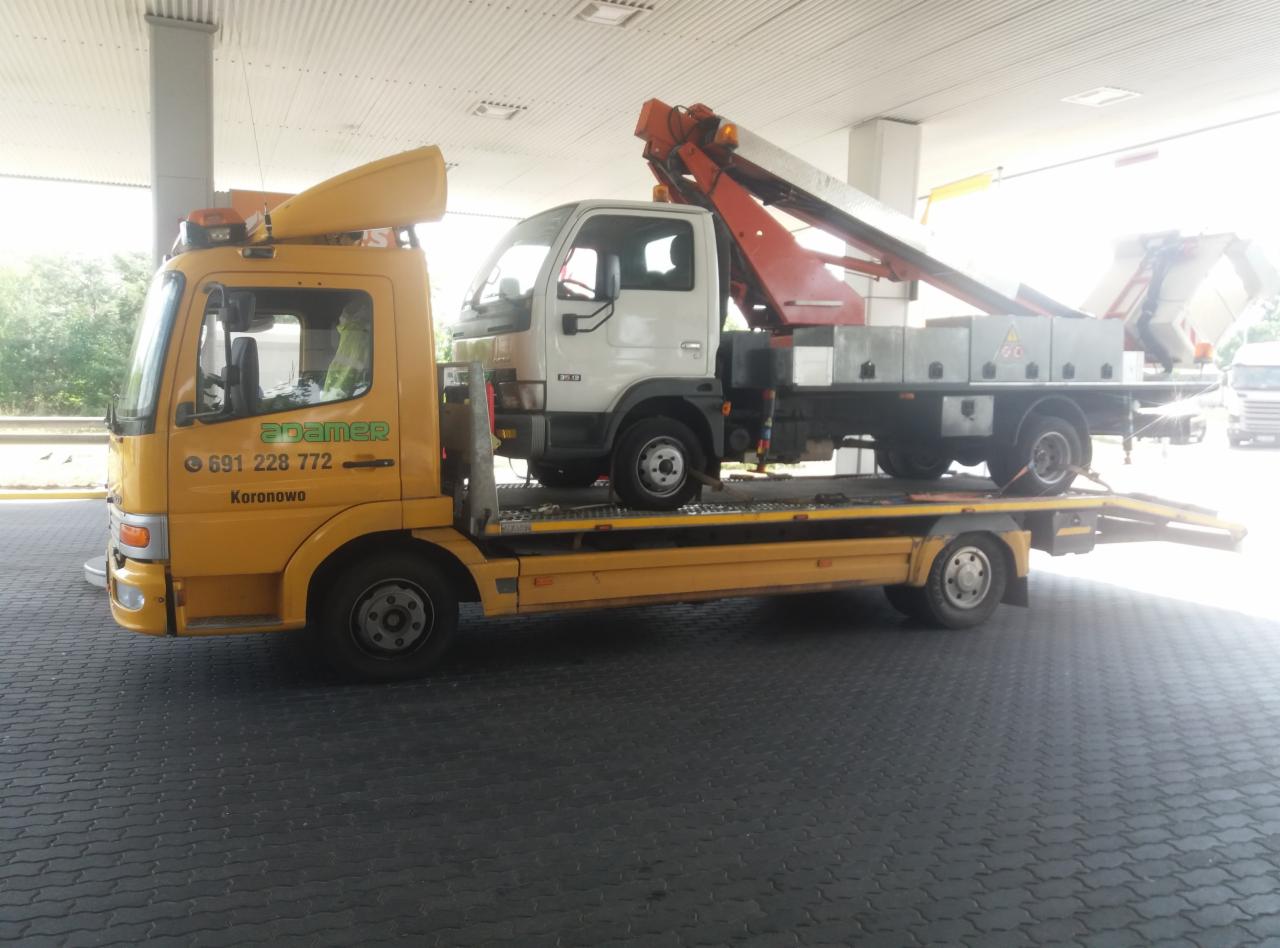 Żółta laweta z białym samochodem na platformie, widoczne logo firmy i numer telefonu na kabinie, zaparkowana pod zadaszeniem stacji benzynowej.