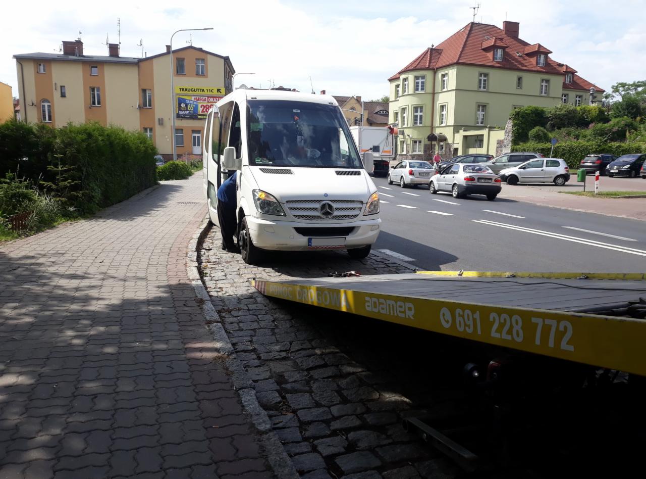 Biały bus Mercedes-Benz Sprinter wciągany na żółtą lawetę na tle miejskiej ulicy z budynkami i reklamą.