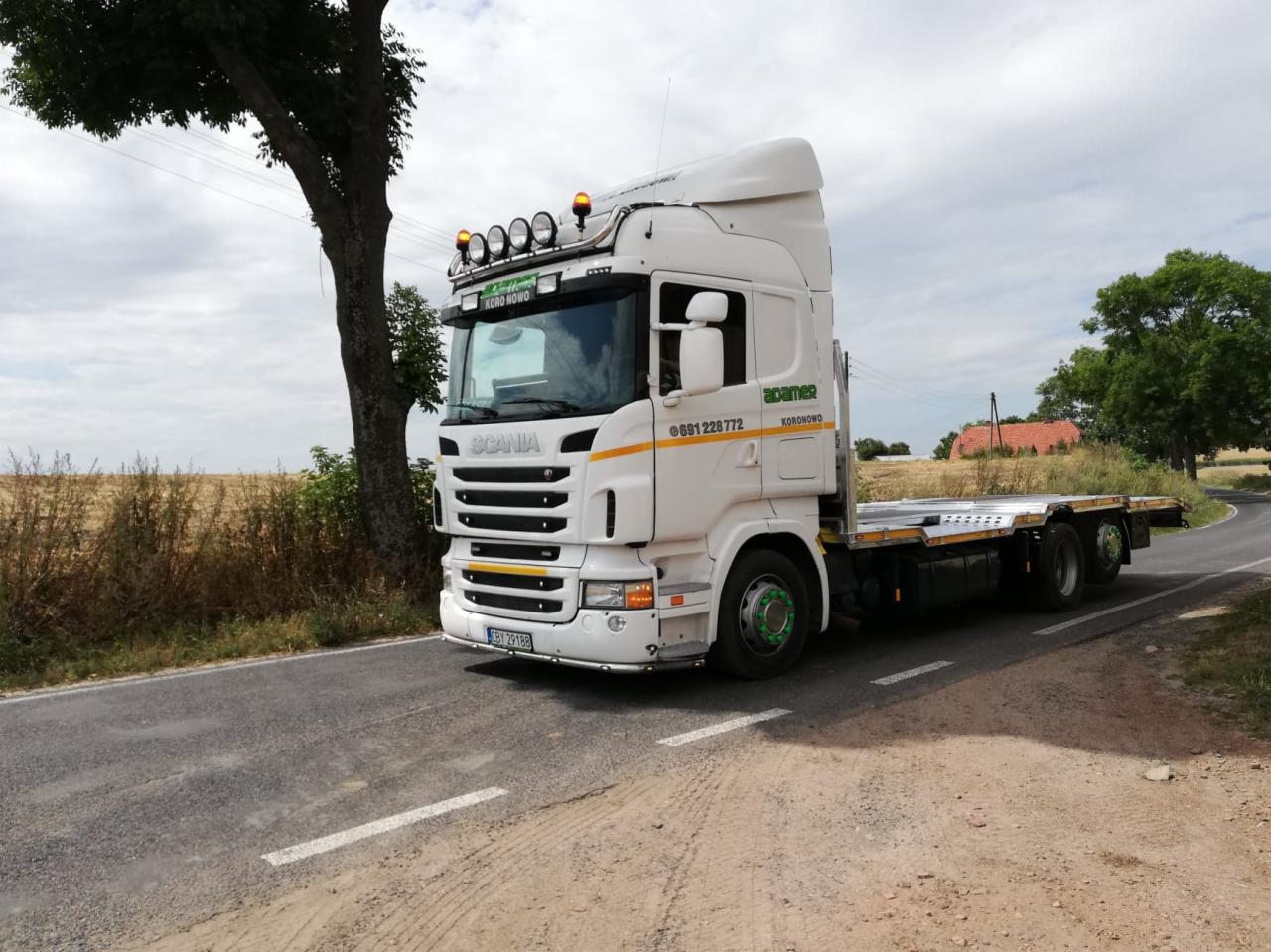 Biała ciężarówka Scania z platformą, zaparkowana na poboczu drogi asfaltowej, z widocznym numerem telefonu i logo firmy, z polami uprawnymi w tle.