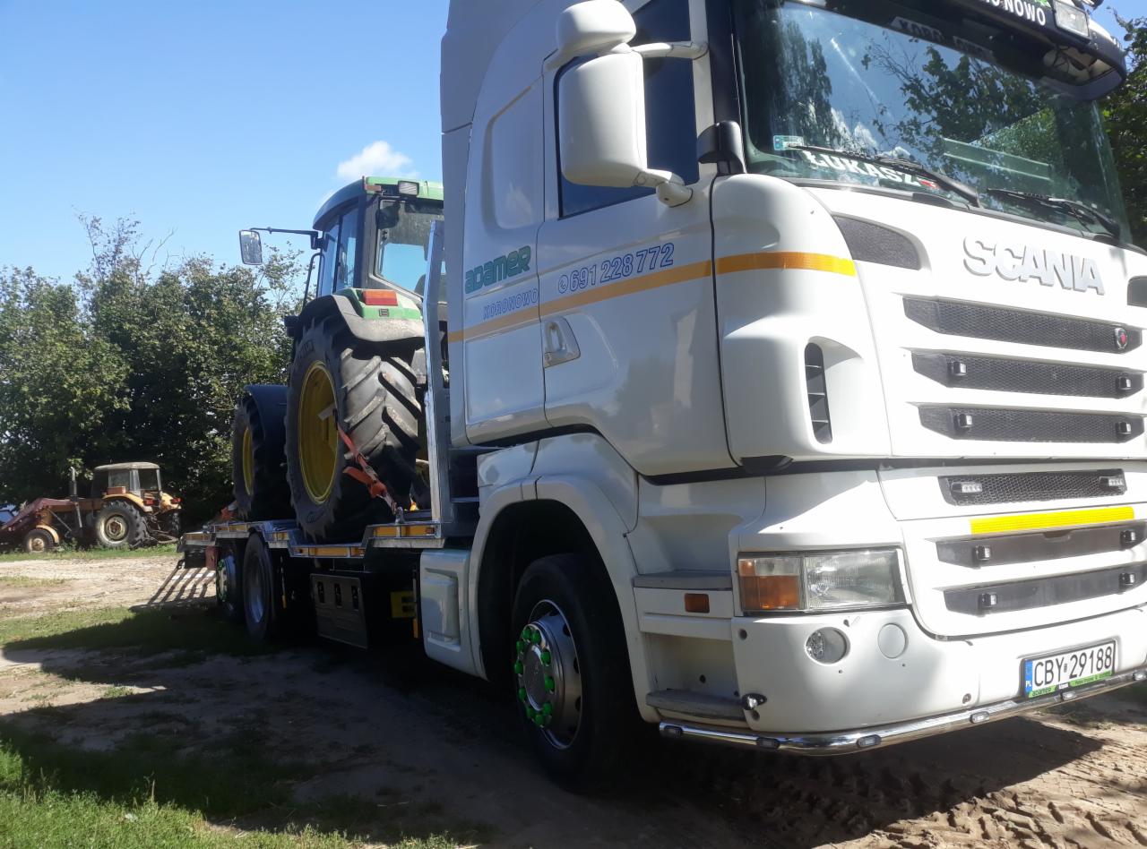 Biała ciężarówka Scania z zielonym ciągnikiem rolniczym na niskopodwoziowej naczepie, widoczne logo firmy transportowej 'adamer' i numer telefonu, w tle kolejny ciągnik.