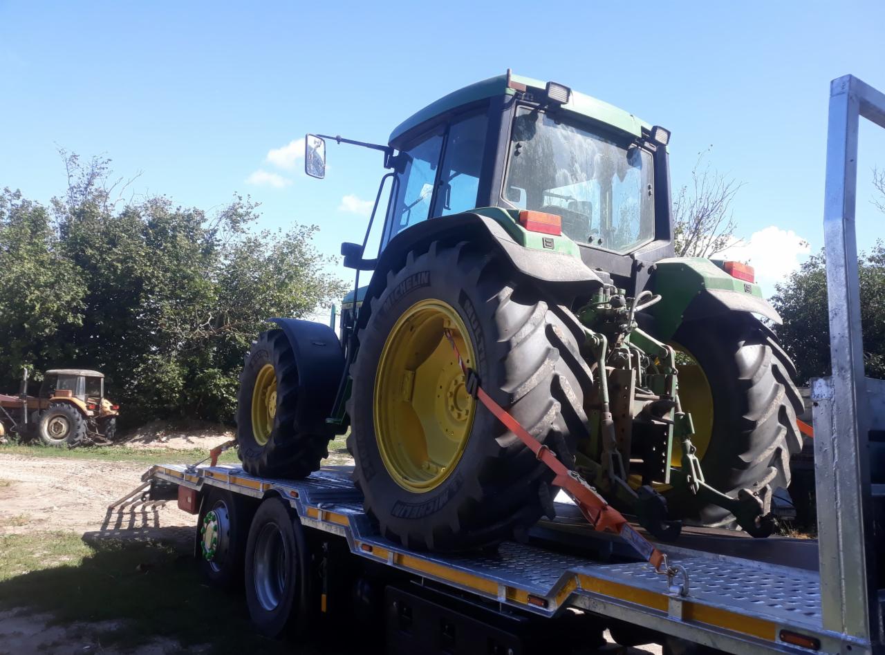 Zielony traktor zabezpieczony pomarańczowymi pasami na platformie niskopodwoziowej, widoczny inny traktor w tle.
