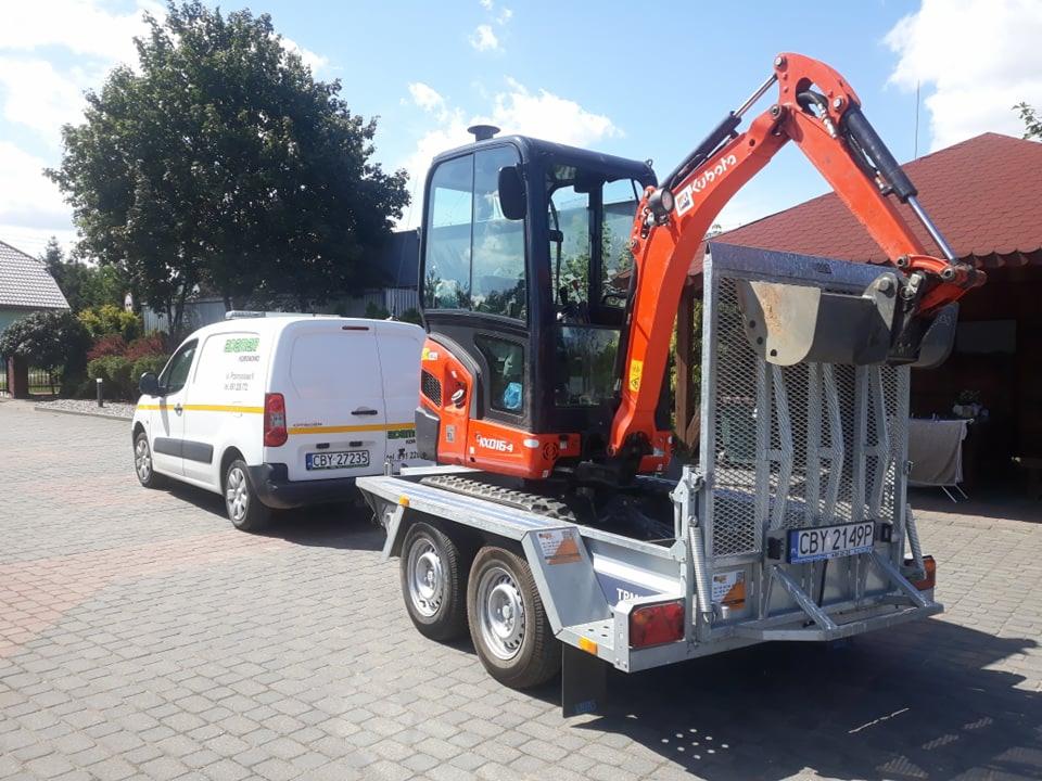 Pomarańczowa mini koparka Kubota na przyczepie, ciągnięta przez biały samochód dostawczy z żółtymi pasami, na brukowanym podwórku.
