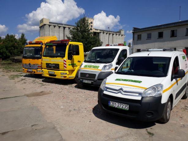 Flota pojazdów pomocy drogowej: dwa żółte samochody ciężarowe MAN i Mercedes oraz dwa białe busy Citroen i Fiat z zielonym logo firmy, zaparkowane na placu z budynkiem w tle.