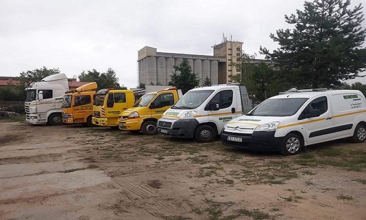 Flota pojazdów firmy transportowej: biała ciężarówka Scania, żółte lawety i busy dostawcze na szutrowym placu, z budynkiem przemysłowym w tle.