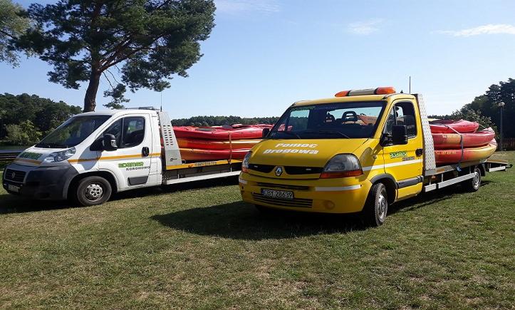 Dwa busy lawety, biały i żółty, zaparkowane na trawie, przewożące czerwone kajaki. Na żółtym busie napis 'Pomoc Drogowa'.