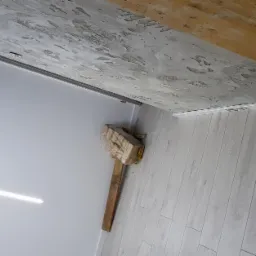 Fragment wnętrza w trakcie prac wykończeniowych: jasne panele podłogowe ułożone, ściana pomalowana na biało z widoczną listwą przypodłogową, sufit z fakturą, zabezpieczony młotek obok ściany...