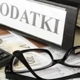 Segregator z napisem 'PODATKI' leżący na stosie dokumentów podatkowych, obok kalkulator, moneta i okulary.