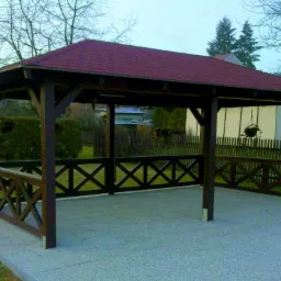 Drewniana altana z czerwonym dachem z gontu bitumicznego, otoczona niskim płotkiem z krzyżowym wzorem, na betonowej wylewce, w tle zielony trawnik i zabudowa jednorodzinna.