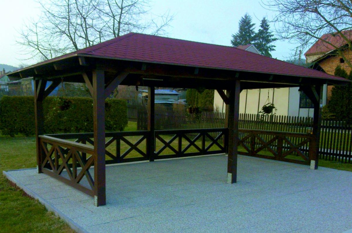 Drewniana altana z czerwonym dachem z gontu bitumicznego, otoczona niskim płotkiem z krzyżowym wzorem, na betonowej wylewce, w tle zielony trawnik i zabudowa jednorodzinna.