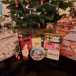 Świąteczny upominek pod choinką: pudełka herbaty, czekoladki Ferrero Rocher, Ptasie Mleczko, Merci, puszka ciastek z motywem zimowym, mała choinka i figurka Świętego Mikołaja.