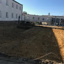 Szeroki wykop w piaszczystej ziemi obok budynku, z widocznymi śladami opon i rurami, koparka w tle.