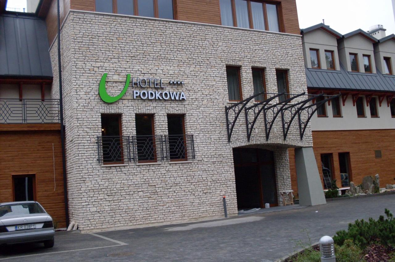 Elewacja budynku hotelu z jasnej cegły z ciemnymi oknami i zielonym logo, widoczny fragment parkingu i samochód osobowy.