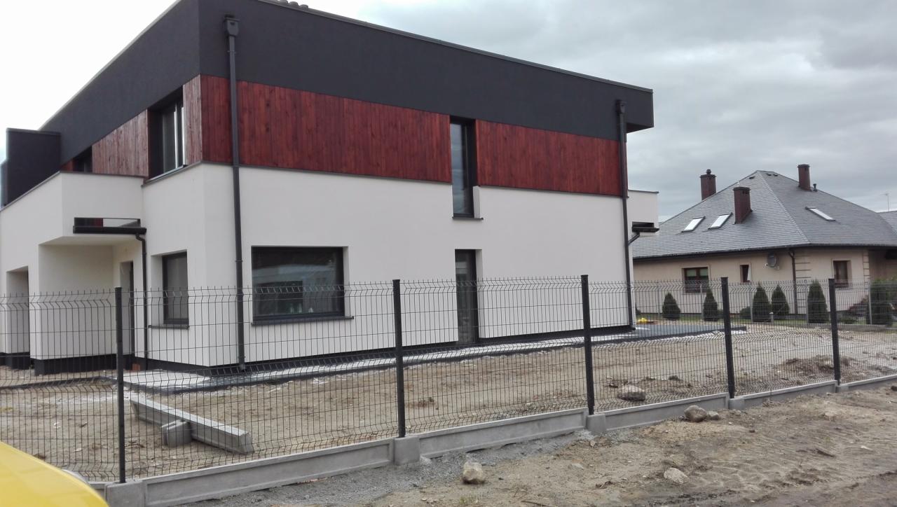 Nowoczesny dom z elementami drewna na elewacji, widoczny z zewnątrz przez metalowe ogrodzenie w trakcie montażu, z drugim domem w tle pod pochmurnym niebem.