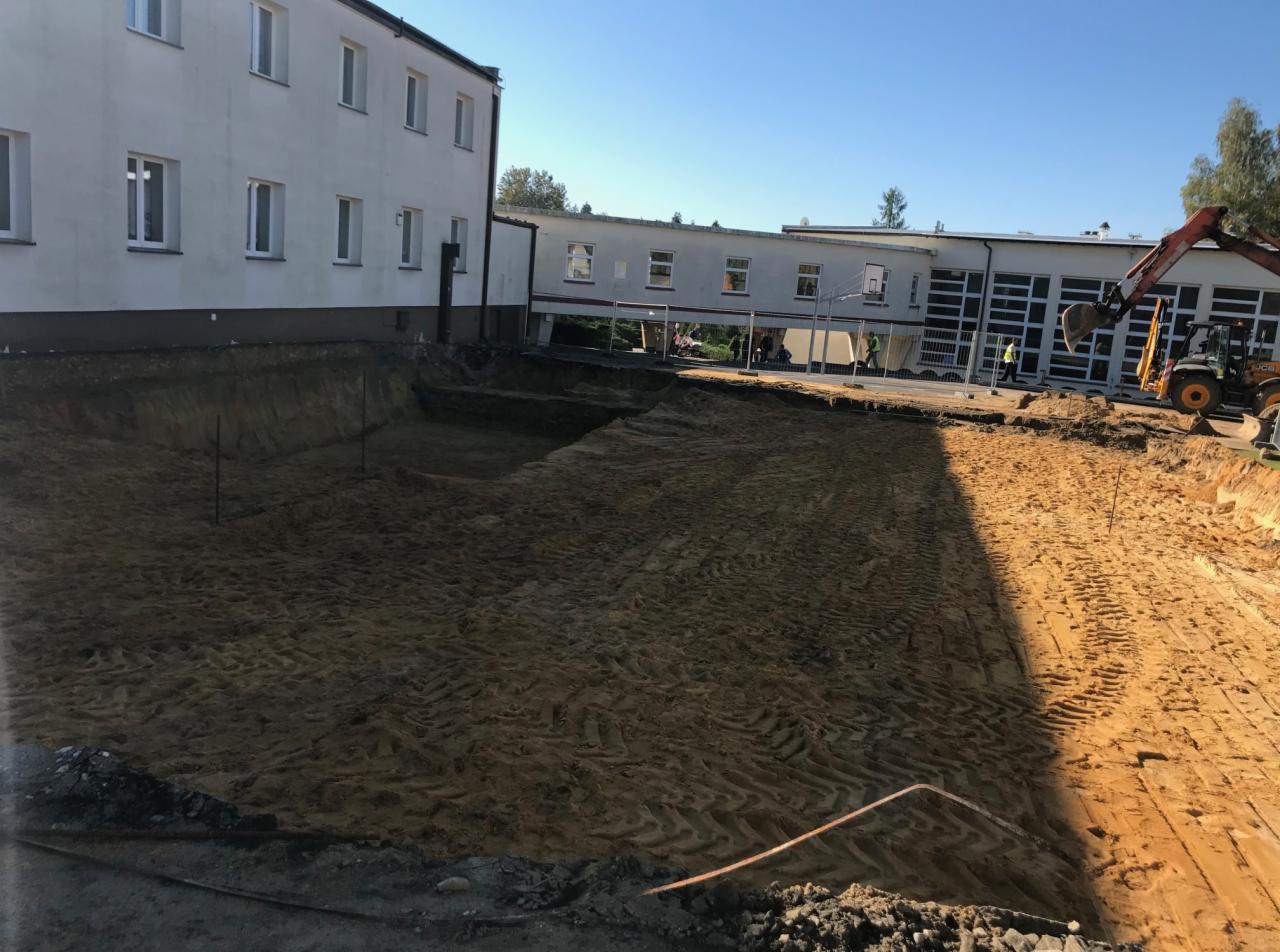 Szeroki wykop w piaszczystej ziemi obok budynku, z widocznymi śladami opon i rurami, koparka w tle.