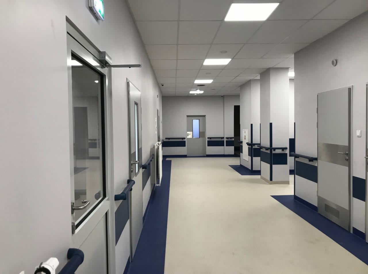 Wnętrze korytarza w budynku użyteczności publicznej z białymi ścianami, szarymi drzwiami, niebieską listwą przypodłogową i poręczami, oraz jasną podłogą.