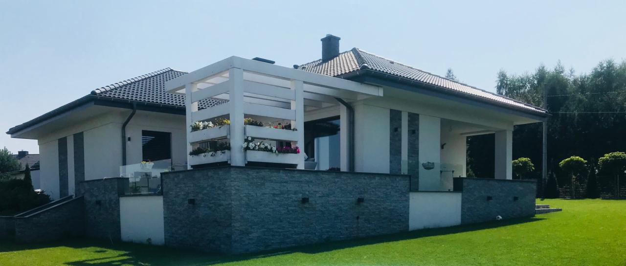 Nowoczesny dom jednorodzinny z białymi ścianami, szarą kamienną elewacją i drewnianą pergolą z kwiatami, otoczony zielonym trawnikiem.