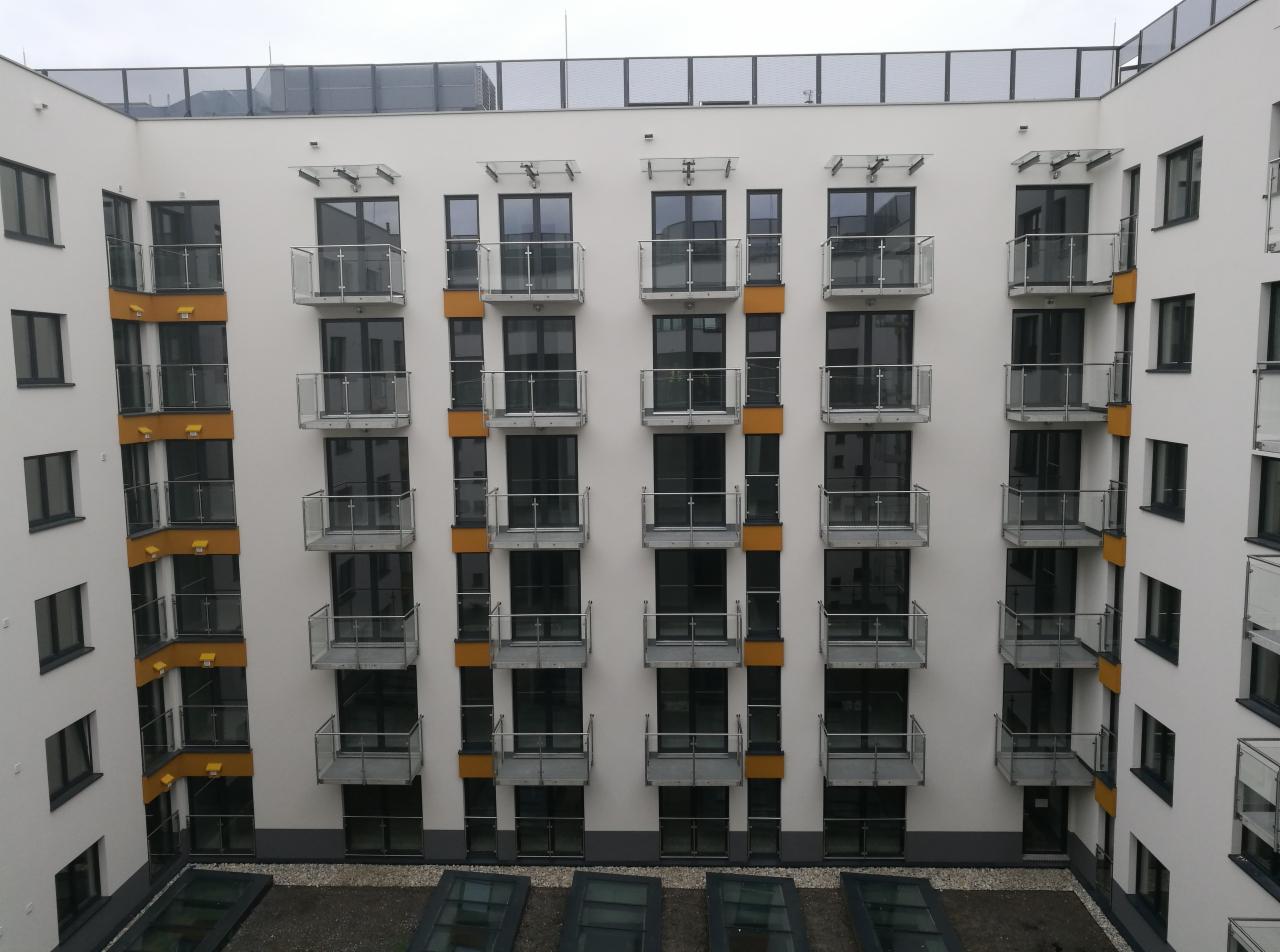 Fasada budynku mieszkalnego z balkonami zabezpieczonymi szklanymi balustradami, widok z góry na wewnętrzny dziedziniec z przeszklonymi elementami.