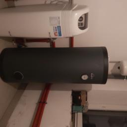 Usługi Hydrauliczne Mateusz Olkowicz - Zamontowany, pionowy, grafitowy zasobnik ciepłej wody użytkowej Kospel z widocznym analogowym termometrem, podwieszony pod białym bojlerem elektrycznym z czerwonymi rurami instalacji grzewczej...