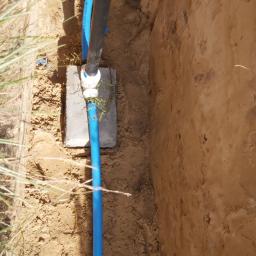 Usługi Hydrauliczne Mateusz Olkowicz - Wykop z widocznymi rurami wodociągowymi, czarna rura pionowa z niebieskim złączem, niebieska rura pionowa na betonowej płycie, instalator w tle.