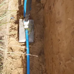 Wykop z widocznymi rurami wodociągowymi, czarna rura pionowa z niebieskim złączem, niebieska rura pionowa na betonowej płycie, instalator w tle.