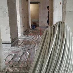 Usługi Hydrauliczne Mateusz Olkowicz - Szpula rur ogrzewania podłogowego na folii z siatką, widok na surową przestrzeń z pustaków, rozłożone pętle rur w tle, instalacja w trakcie realizacji.