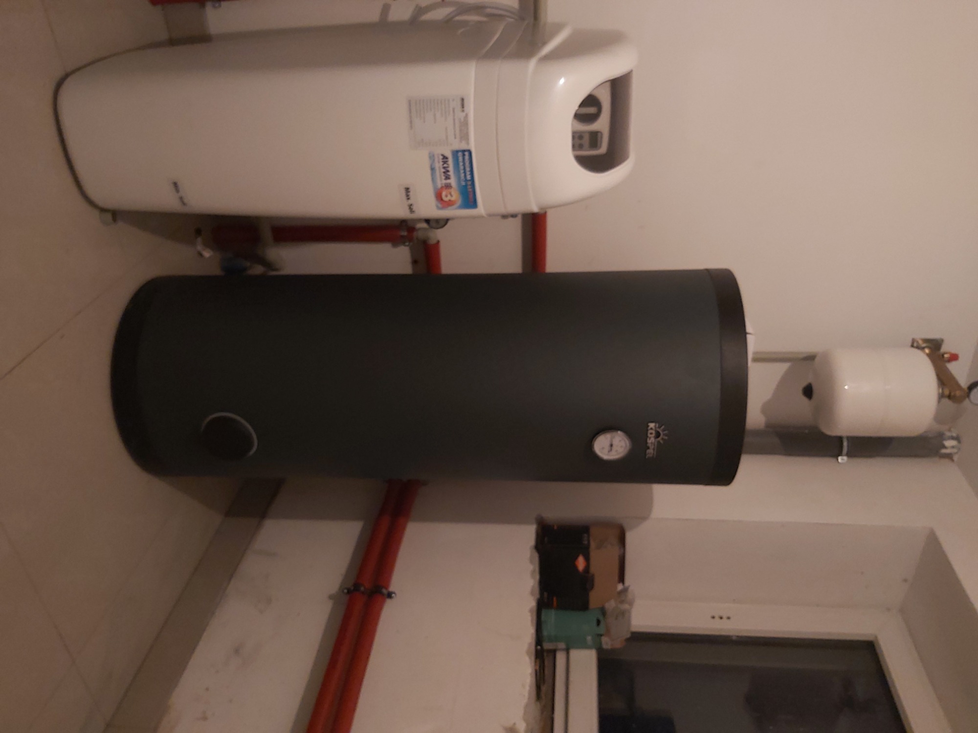 Zamontowany, pionowy, grafitowy zasobnik ciepłej wody użytkowej Kospel z widocznym analogowym termometrem, podwieszony pod białym bojlerem elektrycznym z czerwonymi rurami instalacji grzewczej...