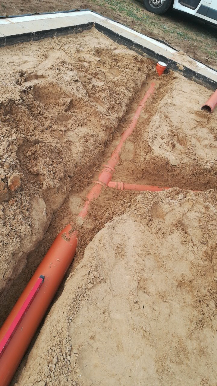 Wykonane wykopy pod fundamenty z widocznymi rurami kanalizacyjnymi w kolorze pomarańczowym, częściowo zasypane piaskiem.