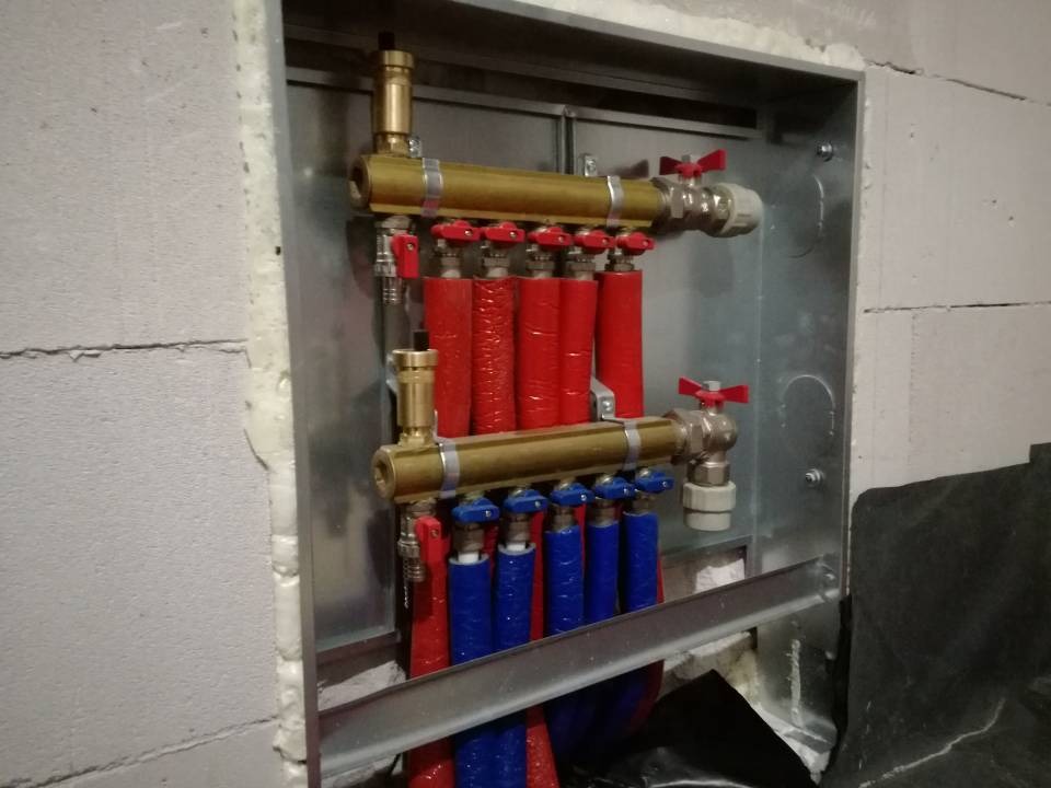 Rozdzielacz ogrzewania podłogowego z czerwonymi i niebieskimi rurami w metalowej szafce, zamontowany w surowej ścianie z betonu komórkowego.