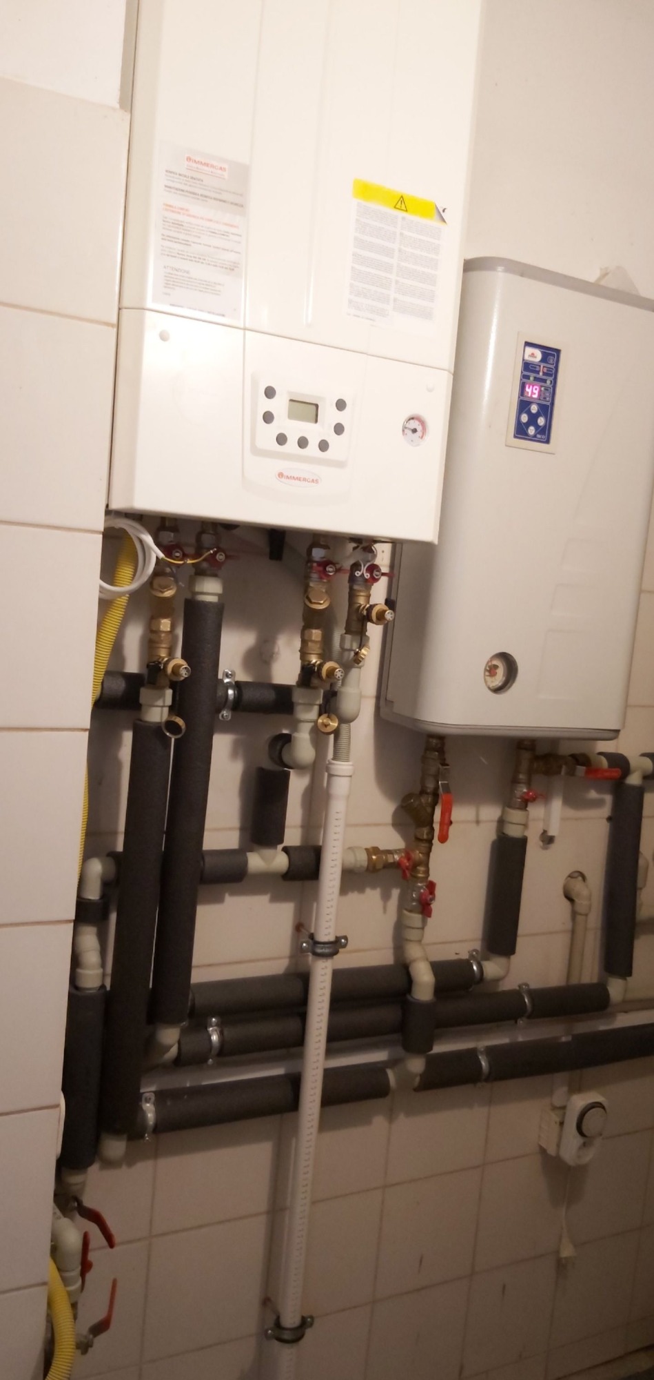 Dwa białe piece, jeden z wyświetlaczem i manometrem, drugi z cyfrowym wyświetlaczem temperatury, połączone rurami miedzianymi i izolowanymi rurami grzewczymi na tle kafelkowanej ściany.