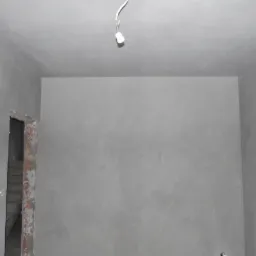 Wnętrze w trakcie prac wykończeniowych: ściany pokryte świeżym, szarym tynkiem, widoczne niedoskonałości na powierzchni, surowa oprawa drzwiowa z ceglanym obramowaniem, wystające przewody elektryczne.