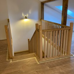 Mariana Podliuk - Drewniane schody z balustradą w jasnym odcieniu drewna, widoczne taśmy malarskie zabezpieczające podłogę, oświetlenie punktowe na ścianie, fragment konstrukcji drewnianej poddasza.