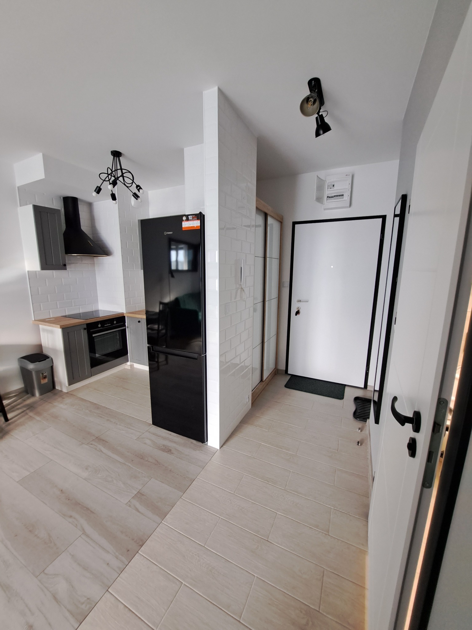 Wnętrze mieszkania po remoncie: widok na kuchnię z szarymi szafkami, czarnym okapem i lodówką, oddzieloną od przedpokoju ścianką z białych płytek, jasne płytki podłogowe imitujące drewno.